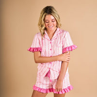 A person smiling and looking down, wearing The Darling Effect’s Oh So Lovely Slumber Party Pajama Shorts Set in pink and white striped ultra-soft fabric with pink ruffles, standing against a beige background.