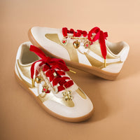 A pair of white and gold sneakers with red velvet laces, adorned with The Darling Effect's Beary in Love Dazzle Shoe Charm Pack and faux pearls, displayed on a beige surface.