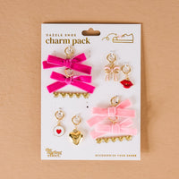 The Darling Effect’s Velvet Crush Dazzle Shoe Charm Pack features six gold and pink Valentine’s charms—bows, hearts, lips, and more—displayed on a white card with a tan background. Perfect for styling your footwear!.
