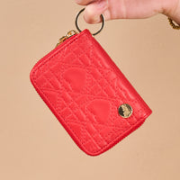 A hand holds a red, quilted Red Hearts Zip Around Wallet by The Darling Effect, featuring a circular gold emblem and an attached keyring.