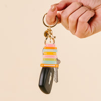 A hand with manicured nails holds a keyring featuring the-darling-effect’s Book Stack Acrylic Keychain, a striped silicone accessory, two silver keys, and a black car key fob against a beige background.