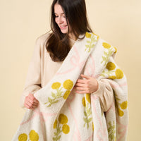 A woman with long brown hair, in a beige sweater, smiles while holding the-darling-effect’s Marigold Lane Pink Cozy Dreams Luxe Blanket—white with yellow, green, and pink floral patterns—against a light beige background.