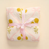 A white towel with yellow polka dots and pink ribbon designs, tied with a light pink bow on a cream background, pairs perfectly with the-darling-effect’s Marigold Lane Pink Cozy Dreams Luxe Blanket for added comfort.