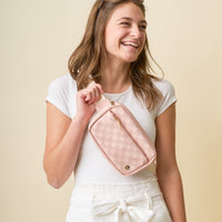A smiling woman in a white t-shirt and high-waisted white pants holds The Darling Effect's Blush Urban Check Belt Bag, a light pink vegan leather accessory, against a plain beige background.
