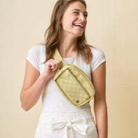 A smiling woman with long brown hair wears a white t-shirt and high-waisted white pants, posing with The Darling Effect's Green Urban Check Belt Bag against a beige background.