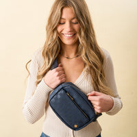 A woman with long, wavy blonde hair smiles while holding The Darling Effect's Navy Urban Check Belt Bag against her beige ribbed top. The background is a plain, light cream color.