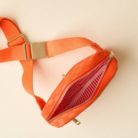 The Darling Effect’s Orange Urban Check Belt Bag features vegan leather, a gold buckle and zipper, and a red-and-white striped lining—shown open against a light beige background for a stylish, versatile accessory.