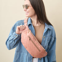 A young woman with long dark hair, sunglasses, and a denim jacket smiles while holding The Darling Effect’s Rose Trendy Luxe Belt Bag in woven pink vegan leather.