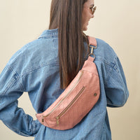 Woman with long brown hair wears sunglasses, a blue denim jacket, and The Darling Effect's Rose Trendy Luxe Belt Bag with gold zippers, posing with her back to the camera against a beige background.