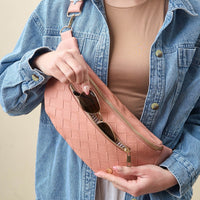 A person in a tan shirt and blue denim jacket puts sunglasses into The Darling Effect’s Rose Trendy Luxe Belt Bag, a pink woven vegan leather crossbody with a gold zipper, highlighting this fashionable accessory.