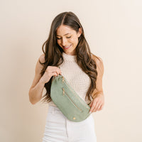 A woman with long brown hair smiles as she unzips The Darling Effect’s Woven Sage Trendy Luxe Belt Bag, styled with a sleeveless knit top and white pants against a plain beige background.