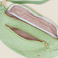 The Darling Effect’s Woven Sage Trendy Luxe Belt Bag features vegan leather in light green with gold zippers, a matching strap for hands-free style, an exterior zip pocket, and is lined with brown and white striped fabric.