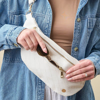 A person in a denim jacket opens The Darling Effect's White Trendy Luxe Belt Bag, revealing sunglasses inside the chic, woven accessory.