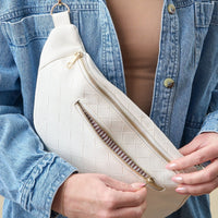 A person in a light blue denim jacket and beige top holds The Darling Effect's White Trendy Luxe Belt Bag, which features vegan leather with a textured pattern and gold zippers, as they open one of its compartments.