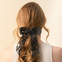 A woman with long, wavy light brown hair is seen from behind in a white sleeveless top, her hair styled with the-darling-effect's Black Ditzy Daisies Hair Bow, adding an elegant touch to her look.