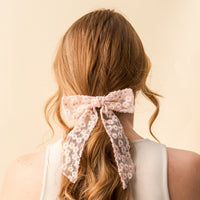 A woman with wavy, light brown hair wears a white top and ties her hair with the Blush Ditzy Daisies Hair Bow from the-darling-effect, featuring a large sheer floral design. The background is soft beige.
