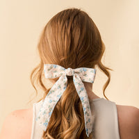 A woman with long, wavy light brown hair tied in a low ponytail with the-darling-effect’s Blue Floral Flair Hair Bow is seen from behind, wearing a sleeveless light top against a plain beige background.