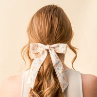 Close-up of a person's long, wavy blonde hair styled with the-darling-effect's Yellow Floral Flair Hair Bow, featuring a white base with yellow flowers, green leaves, and embellished floral details tied at the back.
