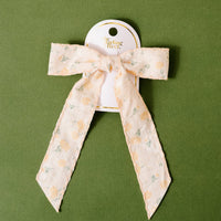 The Yellow Floral Flair Hair Bow by the-darling-effect has embellished floral details and a delicate yellow pattern, with two long tails. Displayed on a white card against a textured green background.