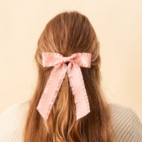 A person with long, wavy light brown hair wears The Darling Effect's Blush Ruffle Hair Bow at the back of their head. Set against a soft beige background, they pair the elegant accessory with a cream sweater.