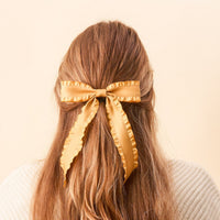 A person with long, light brown hair, seen from behind, wears The Darling Effect’s Mustard Ruffle Hair Bow—a large satin bow with ruffled edges—standing out against a soft beige background.