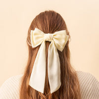 A Cream Satin Hair Bow from The Darling Effect is tied at the back of a person's auburn hair.
