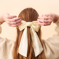 A person's back is shown, wearing a cream sweater and an elegant Cream Satin Hair Bow from The Darling Effect, tied in their auburn hair. Their hands gently hold the accessory on either side of the bow.