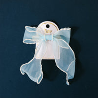 The Blue Sheer Pearl Hair Bow by the-darling-effect features a sheer light blue bow with white trim on a gold barrette, displayed on a white card with gold text against a dark blue background.
