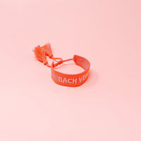 The Darling Effect's Bach Vibes Woven Bracelet features pink adjustable weaving, playful tassels, and "BACH VIBE" in white embroidery. Shown on a pink background, it's perfect for stacking with your favorite trendy bracelets.