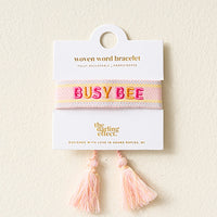 The Darling Effect’s Busy Bee Woven Bracelet features pink and yellow stripes, embroidered "BUSY BEE" text, adjustable fit, tassels, and comes on a display card. Part of The Darling Effect embroidered bracelets collection.
