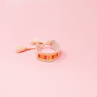 The Darling Effect's Busy Bee Woven Bracelet, shown on a pink backdrop, features "BUSY BEE" embroidered in red and yellow with tassel ends—an adorable, adjustable addition to any bracelet collection.