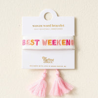 A woven white bracelet with the words BEST WEEKEND in colorful letters, displayed on a card with pink tassels hanging below. The card reads woven word bracelet and the darling effect..