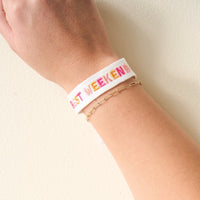 A persons wrist wearing a white fabric bracelet with BEST WEEKEND in pink and yellow letters, alongside a delicate gold chain bracelet.