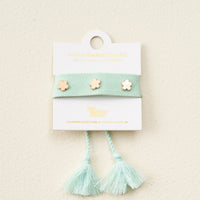 The Darling Effect's Aqua Woven Stacker Bracelet features three gold flower charms, adjustable tassel ends, and is displayed on a white card with gold text.