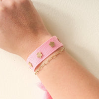 A wrist features The Darling Effect's Pink Woven Bracelet with gold clover-shaped flower charms, stacked with a thin gold chain bracelet—ideal for chic floral stacks against a light background.