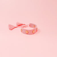 The Darling Effect's Pink Woven Bracelet features gold flower details and two pink tassels—an adjustable accessory ideal for floral stacker styles, shown here on a pink background.