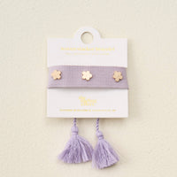 The Darling Effect’s Periwinkle Woven Stacker Bracelet features two tassels and three gold flower charms, displayed on a white card with gold text.