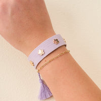 A wrist with two adjustable bracelets: The Darling Effect's Periwinkle Woven Stacker Bracelet—a wide lavender band with flower details and a purple tassel—plus a thin gold chain bracelet, shown against a light beige background.