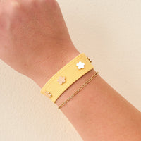 A hand wears The Darling Effect’s Yellow Woven Stacker Bracelet with gold flower detail and a thin gold chain, ideal for creating your own floral stack, set against a light beige background.