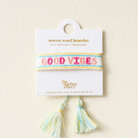 The Darling Effect’s Good Vibes Woven Bracelet features pastel blue, yellow, and white stripes with “GOOD VIBES” in pink embroidery, adjustable fit, tassels at the bottom, and comes displayed on a white card.