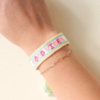 The Darling Effect's Good Vibes Woven Bracelet features white adjustable fabric with "GOOD VIBES" embroidered in pink and light blue—perfect for stacking with a gold chain for a trendy look.