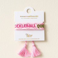 The Darling Effect's Pickleball Queen Woven Bracelet features pink embroidery and tassels, is fully adjustable, and comes on a white card. Designed in Grand Rapids, MI—perfect for fans of trendy embroidered bracelets.