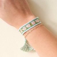 A wrist with two bracelets: one is The Darling Effect’s adjustable "Pickleball Yall" woven bracelet with a tassel, and the other is a thin gold chain featuring small links.