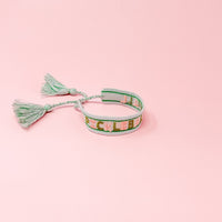 The Darling Effect's Pickleball Yall Woven Bracelet features green, pink, and white threads, "PICKLEBALL YALL" embroidery, and two tassels at the ends, displayed on a light pink background.