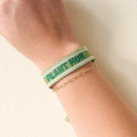 A person’s wrist adorned with The Darling Effect’s Plant Mom Woven Bracelet in green and a delicate gold beaded chain—stylish accessories highlighted on a pale, neutral background.