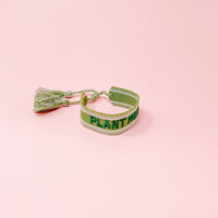 The Darling Effect's Plant Mom Woven Bracelet features "PLANT" in green on a green and white adjustable band with tassels, shown on a pastel pink background. Ideal for trendy accessory lovers and fans of unique embroidered bracelets.