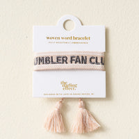 The Darling Effect’s Tumbler Fan Club Woven Bracelet is a trendy, adjustable piece featuring “TUMBLER FAN CLUB” in black, two pink tassels, and comes displayed on a branded card.