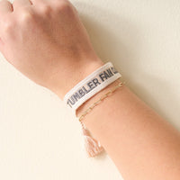 A hand wearing The Darling Effect's Tumbler Fan Club Woven Bracelet with "TUMBLER FAN CLUB" in black embroidery and a trendy gold chain bracelet with tassel, set against a light background.