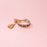 The Darling Effect's Tumbler Fan Club Woven Bracelet features trendy embroidery, playful tassels, and an adjustable fit—perfect for adding style to any outfit. Shown against a pink background.
