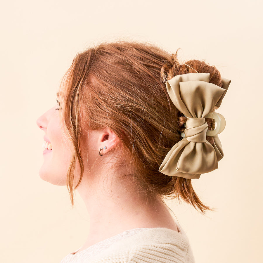A woman with red hair in an updo wears The Darling Effect Green Satin Bow Claw Clip. She smiles facing left against a cream background, styling the chic hair accessory with a white knit top and small hoop earrings.